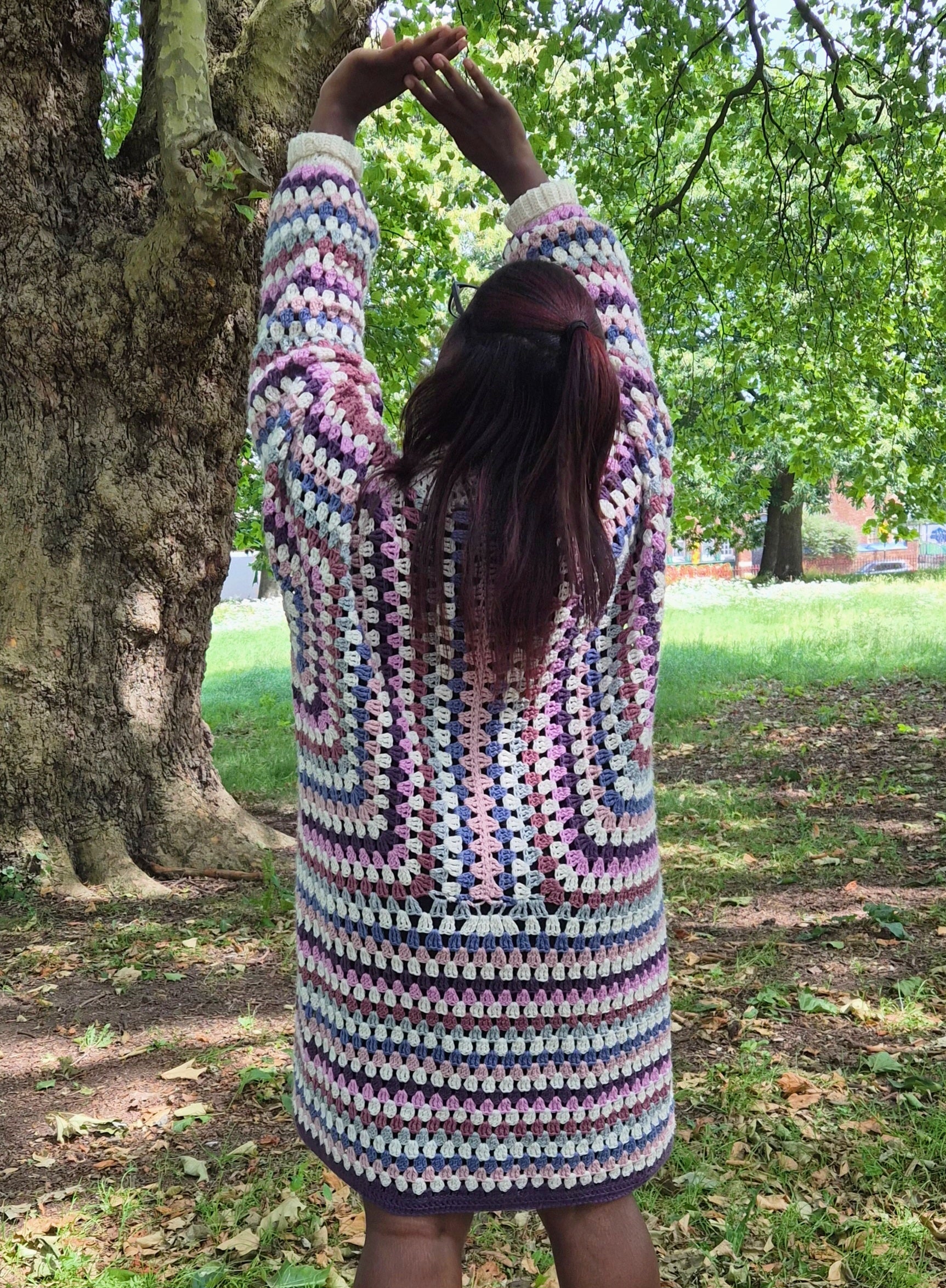A person standing outdoors in a park, viewed from behind, wearing a chunky crocheted cardigan with a multicolor geometric pattern in pink, blue, purple, and white.