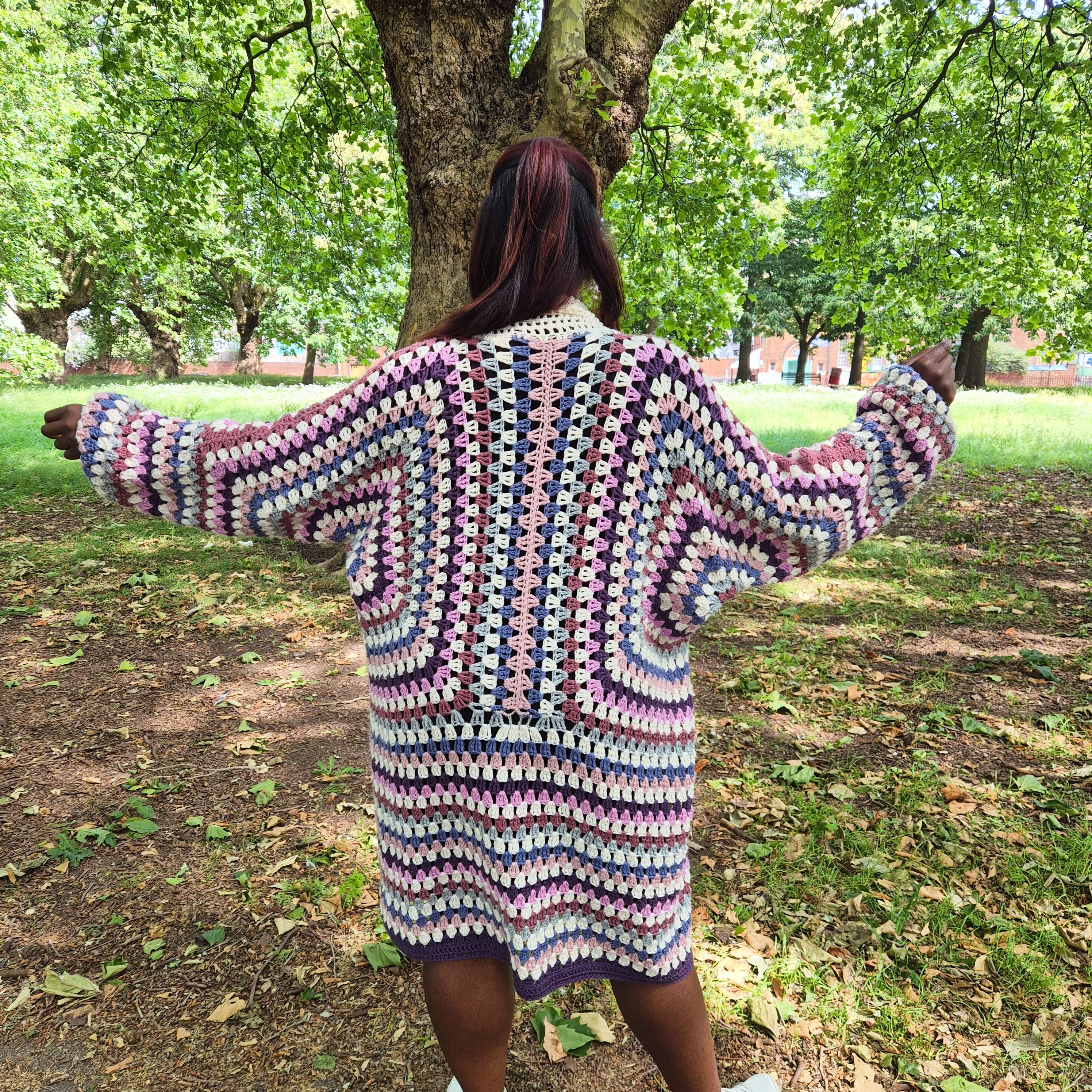A person viewed from behind wearing a chunky, multicolor crochet cardigan with a geometric pattern, standing in a park with trees and grass in the background.