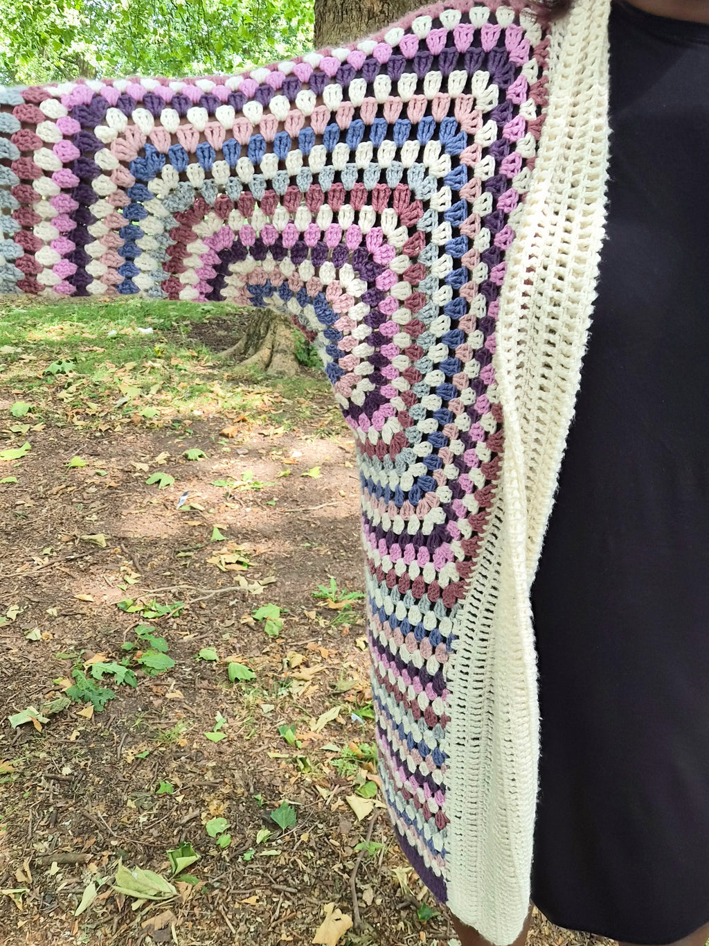 A multicolor crocheted cardigan with concentric rainbow stripes and a cream-colored lace trim, worn by a person outdoors in a wooded area.