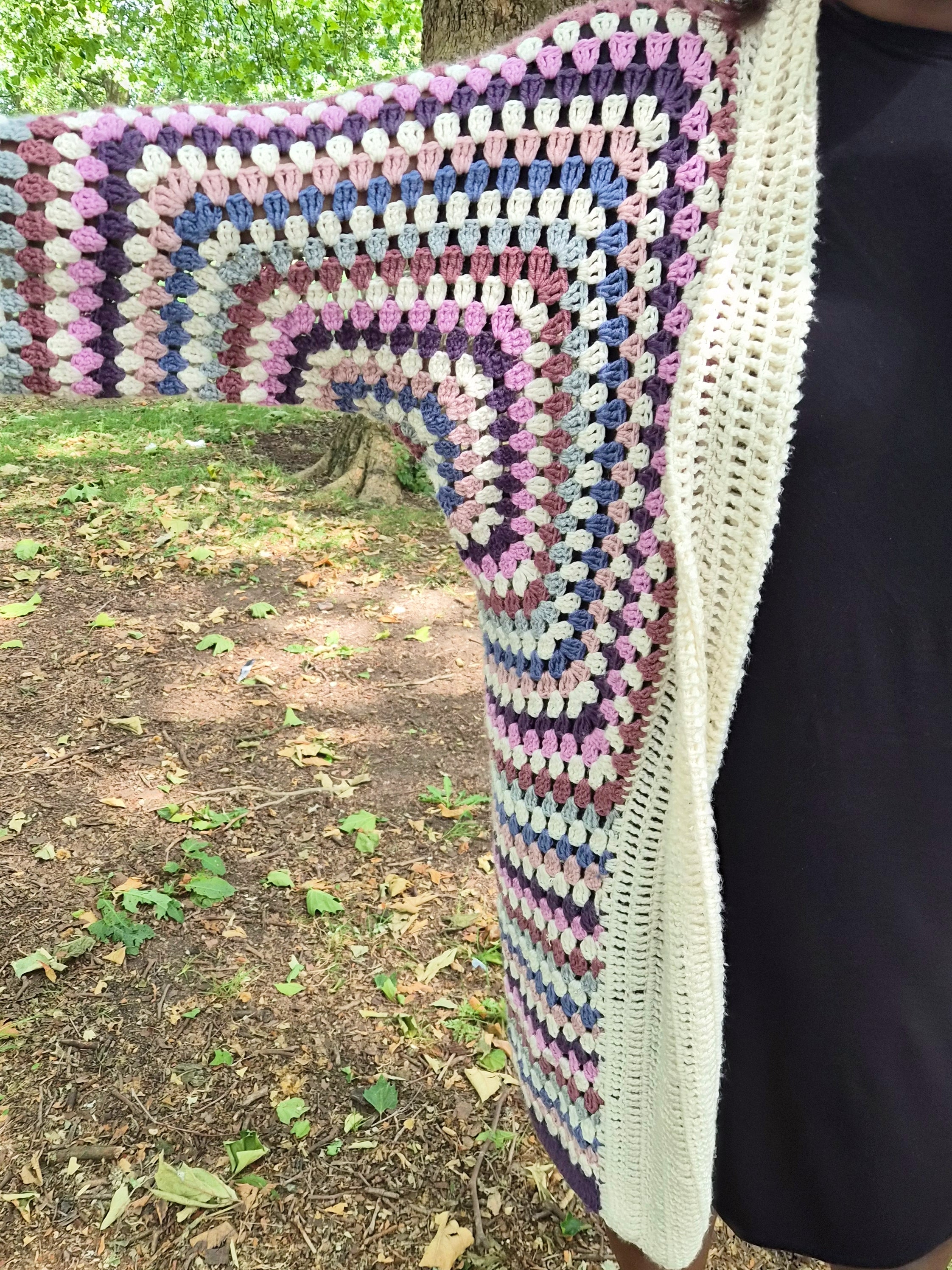 A multicolor crocheted cardigan with concentric rainbow stripes and a cream-colored lace trim, worn by a person outdoors in a wooded area.