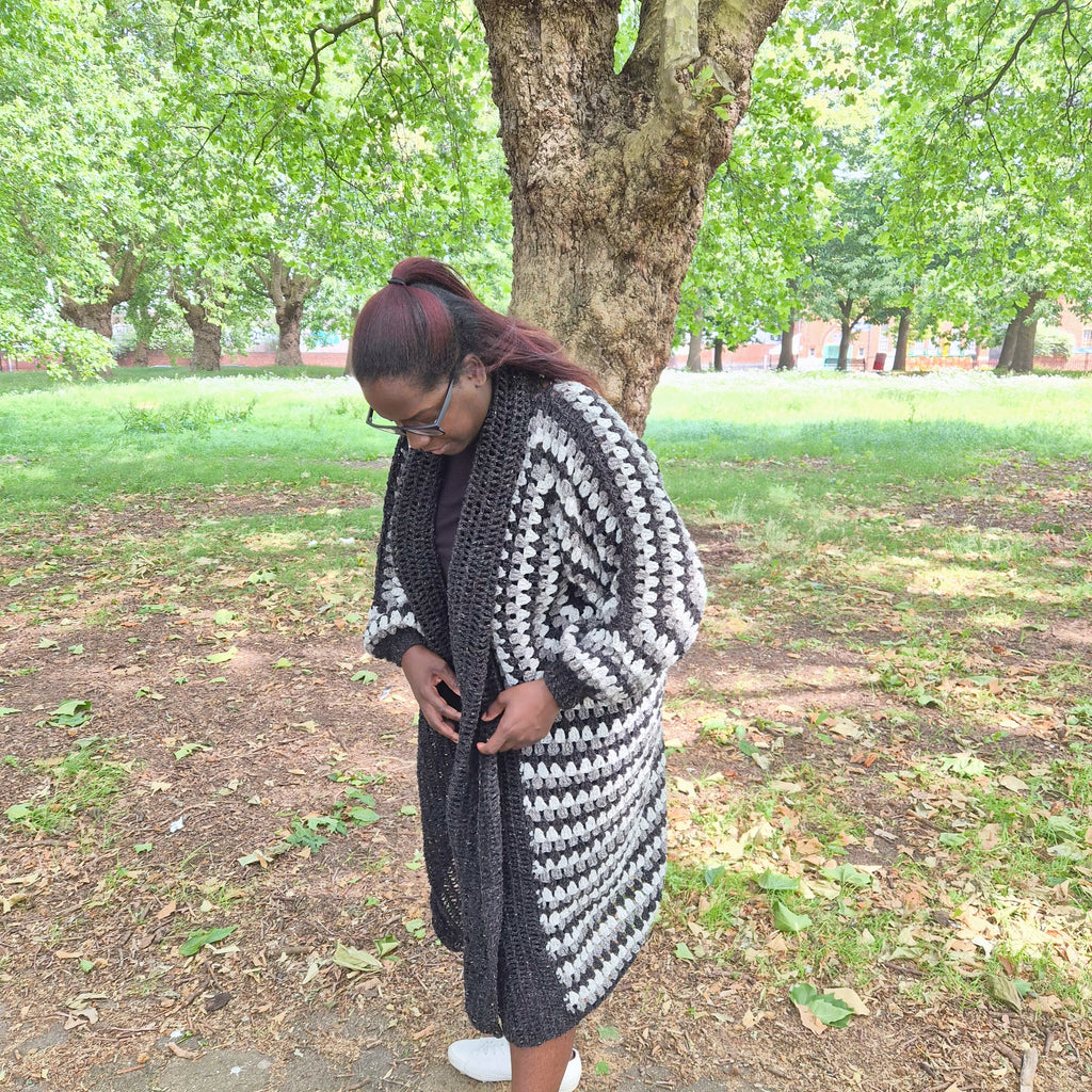A woman wearing a long, chunky black and white crocheted cardigan and a matching black dress, standing outdoors in a park with a large tree behind her.