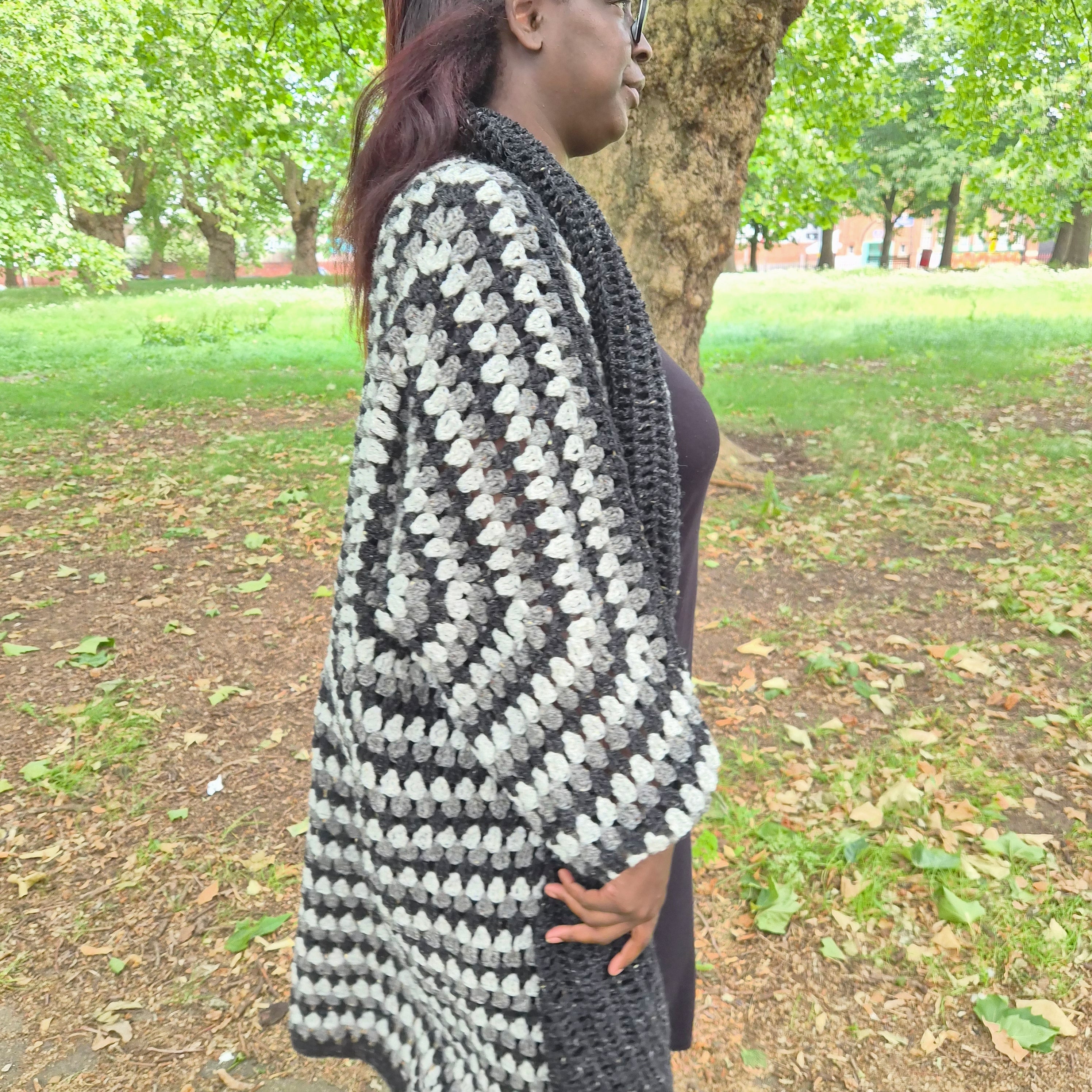 A woman wearing a chunky black and white crochet wrap cardigan in an outdoor park setting with trees and grass in the background.