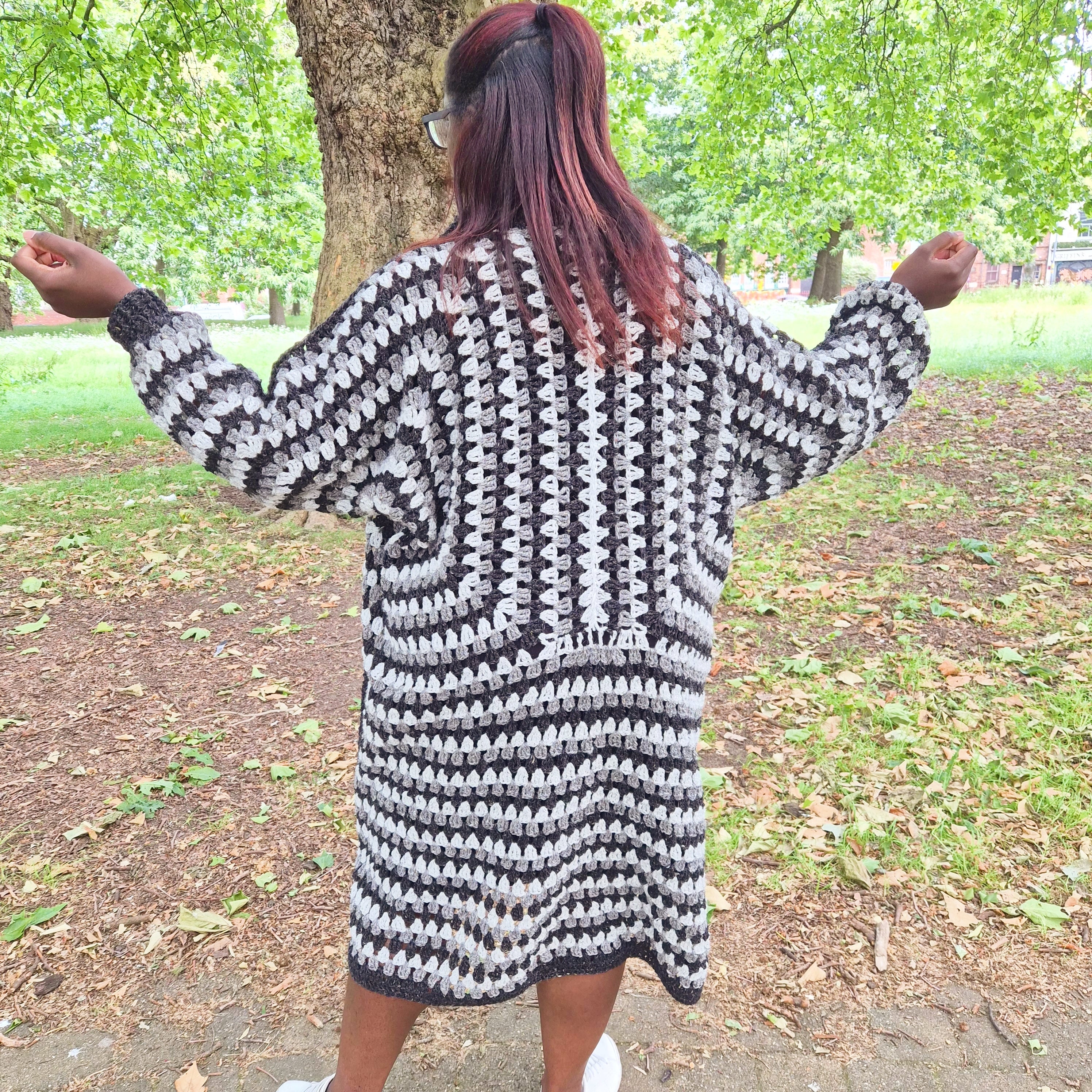 A person standing outdoors in a park wearing a chunky black and white crochet cardigan, viewed from behind with arms outstretched.