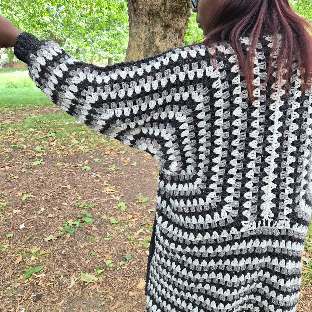 A person outdoors wearing a chunky black and white crocheted cardigan, photographed from the back and side. The cardigan has a textured, granny-square style pattern with alternating black and white yarn.