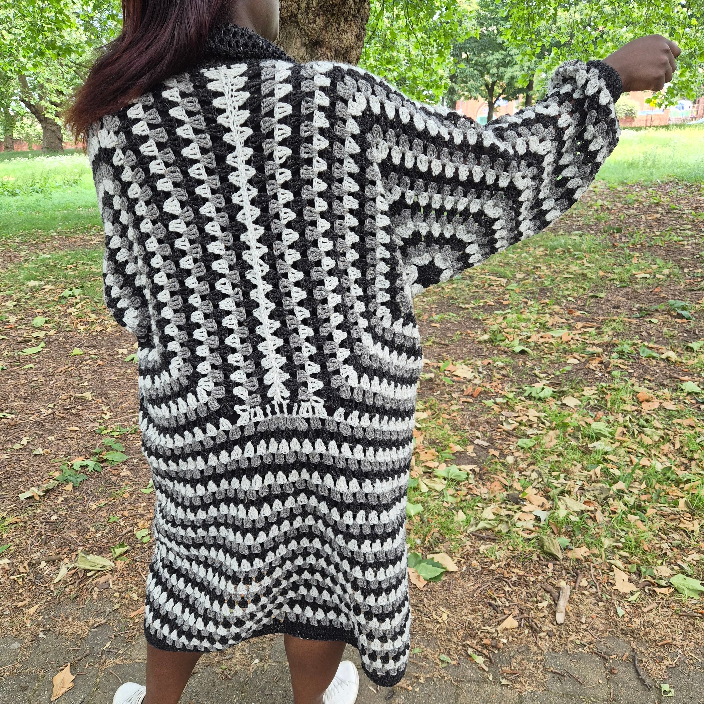 A person standing outdoors in a park wearing a black and white crocheted cardigan or sweater dress, photographed from the back.