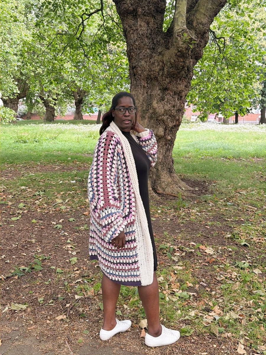 A woman wearing a long, open-front chunky crochet cardigan with a geometric pattern in cream, burgundy, and gray, styled over a black dress and white sneakers, standing in a park with a large tree behind her.