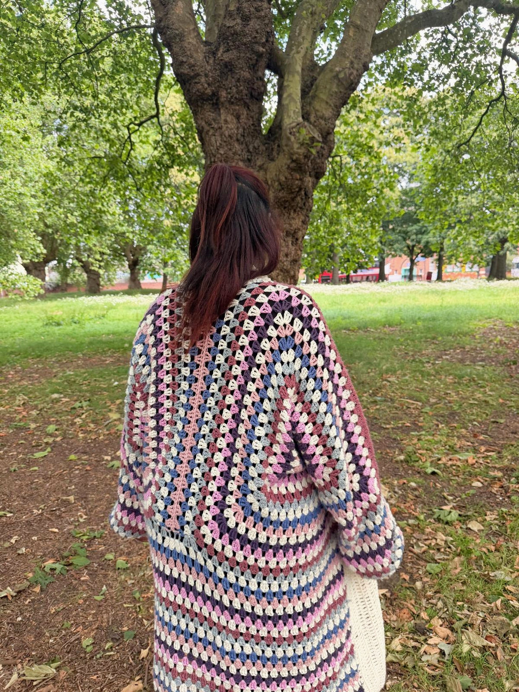 A person standing outdoors in a park, viewed from behind, wearing a chunky crocheted cardigan with a multicolor spiral pattern of pink, blue, purple, and cream.