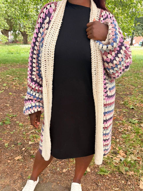 A person outdoors wearing a chunky crocheted cardigan with a multicolor granny-square pattern in pink, blue, purple, and cream, layered over a black dress and white sneakers.