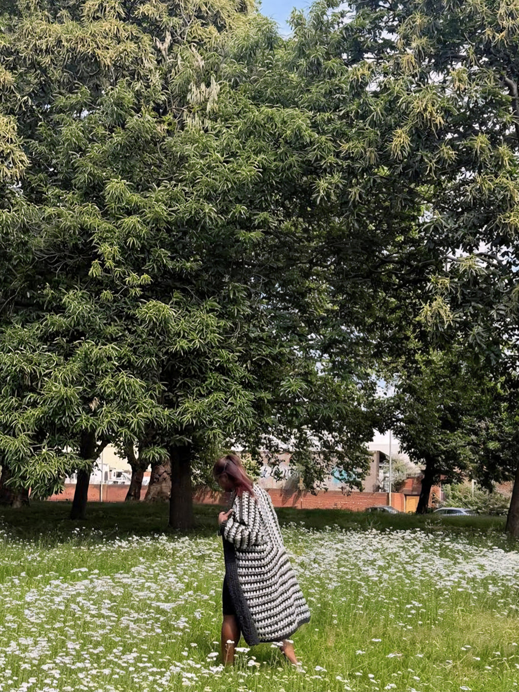 A woman stands in a grassy field of small white flowers under large green trees, wearing a long black-and-white patterned cardigan over a dark dress.