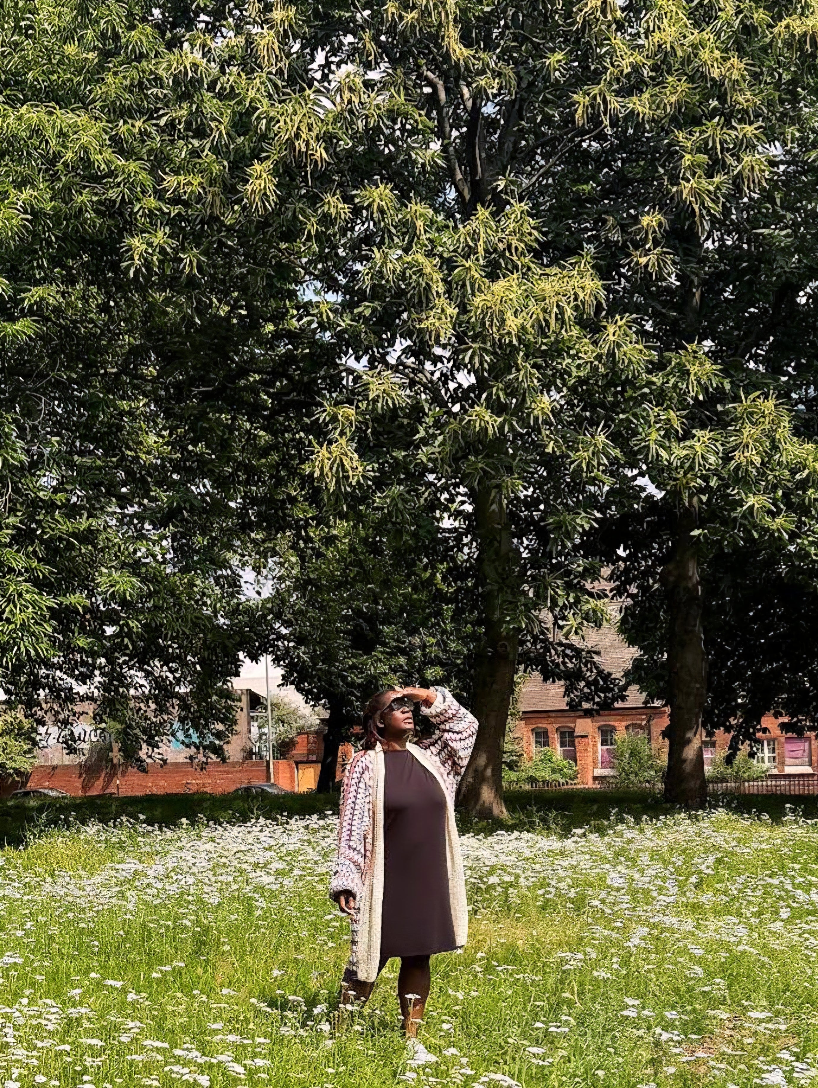A woman stands in a grassy field with white flowers under large green trees, wearing a black dress, a multi coloured crocheted cardigan, and white sneakers