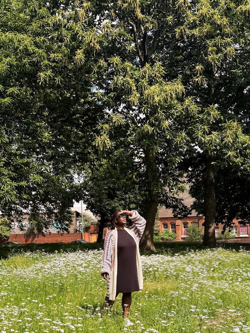 A woman stands in a grassy field with white flowers under large green trees, wearing a black dress, a multi coloured crocheted cardigan, and white sneakers